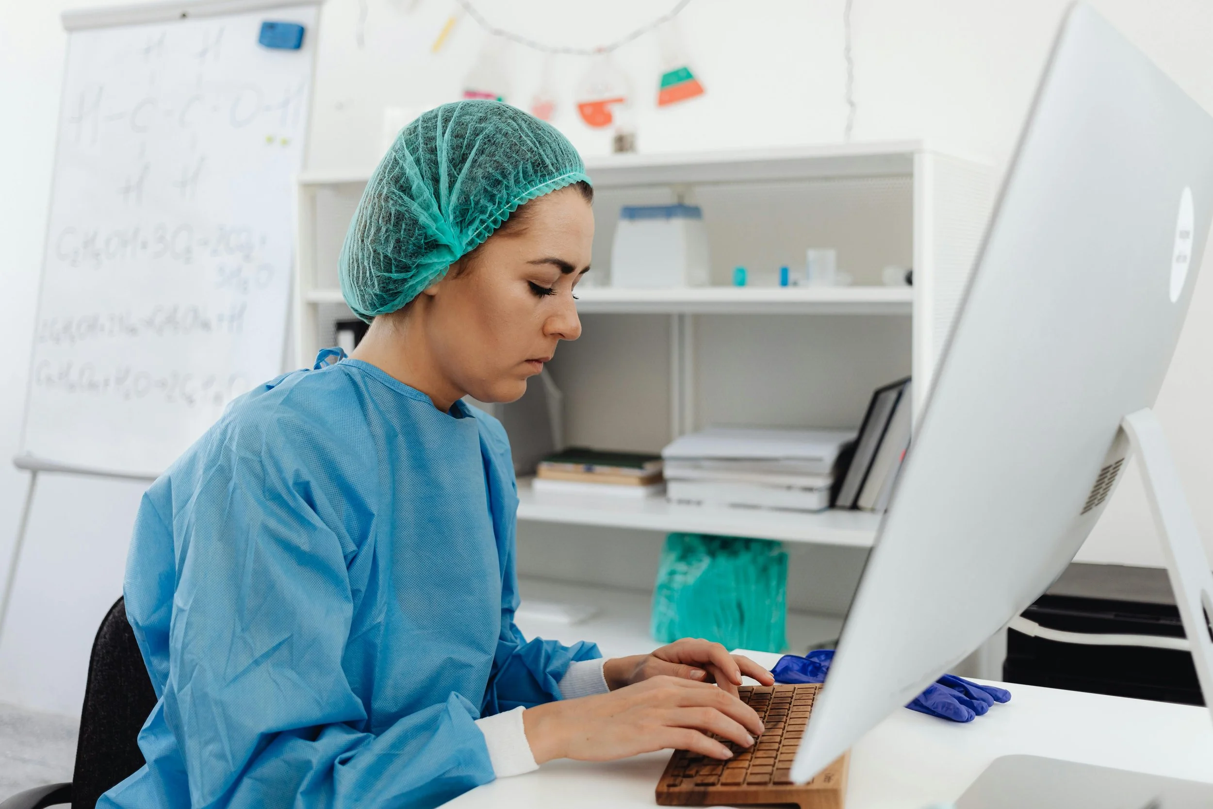 A clinician in scrubs and a surgical cap typing at a desktop computer