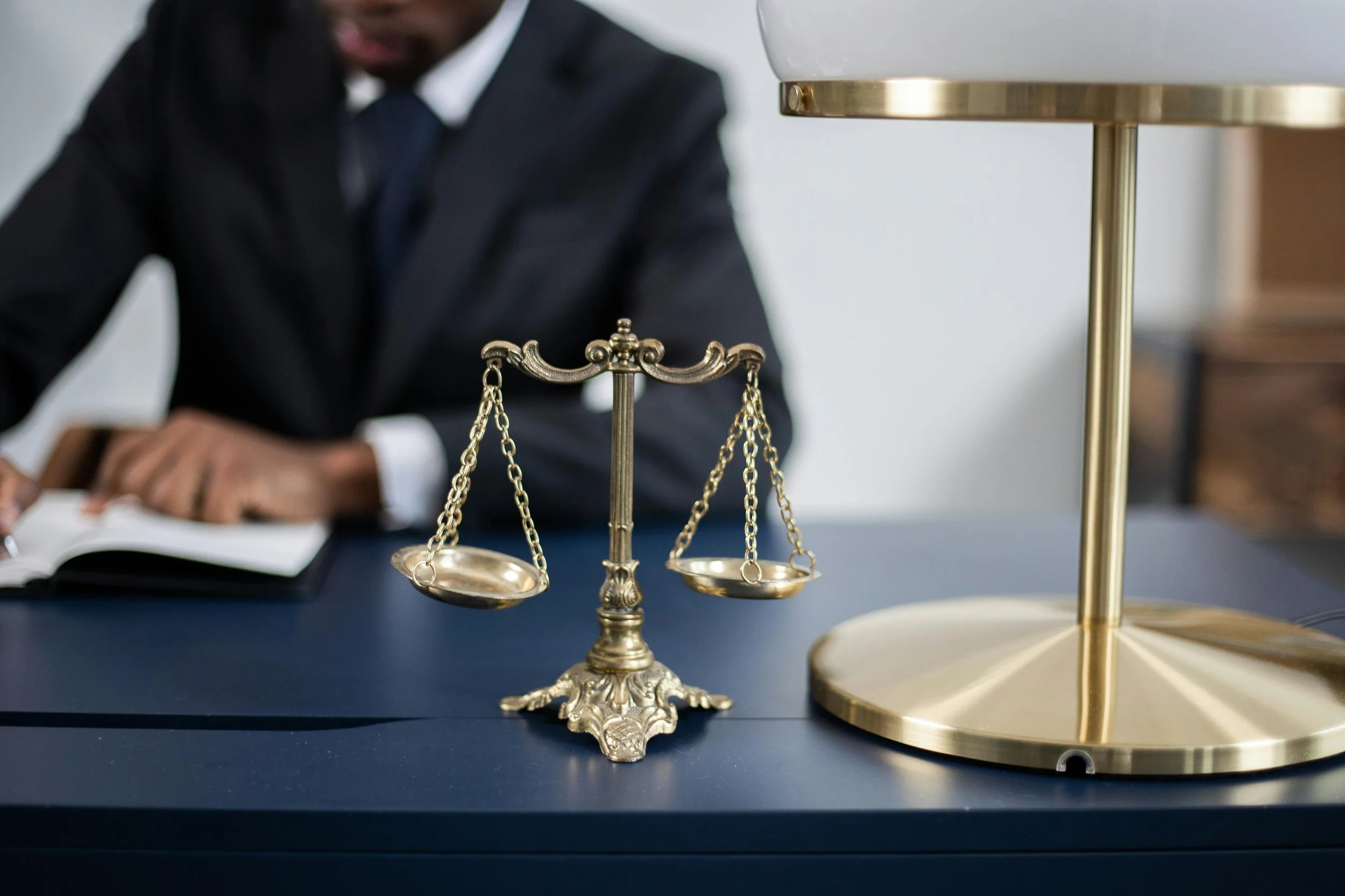 Brass scales of justice on a desk while a person in a suit reads a document in the background