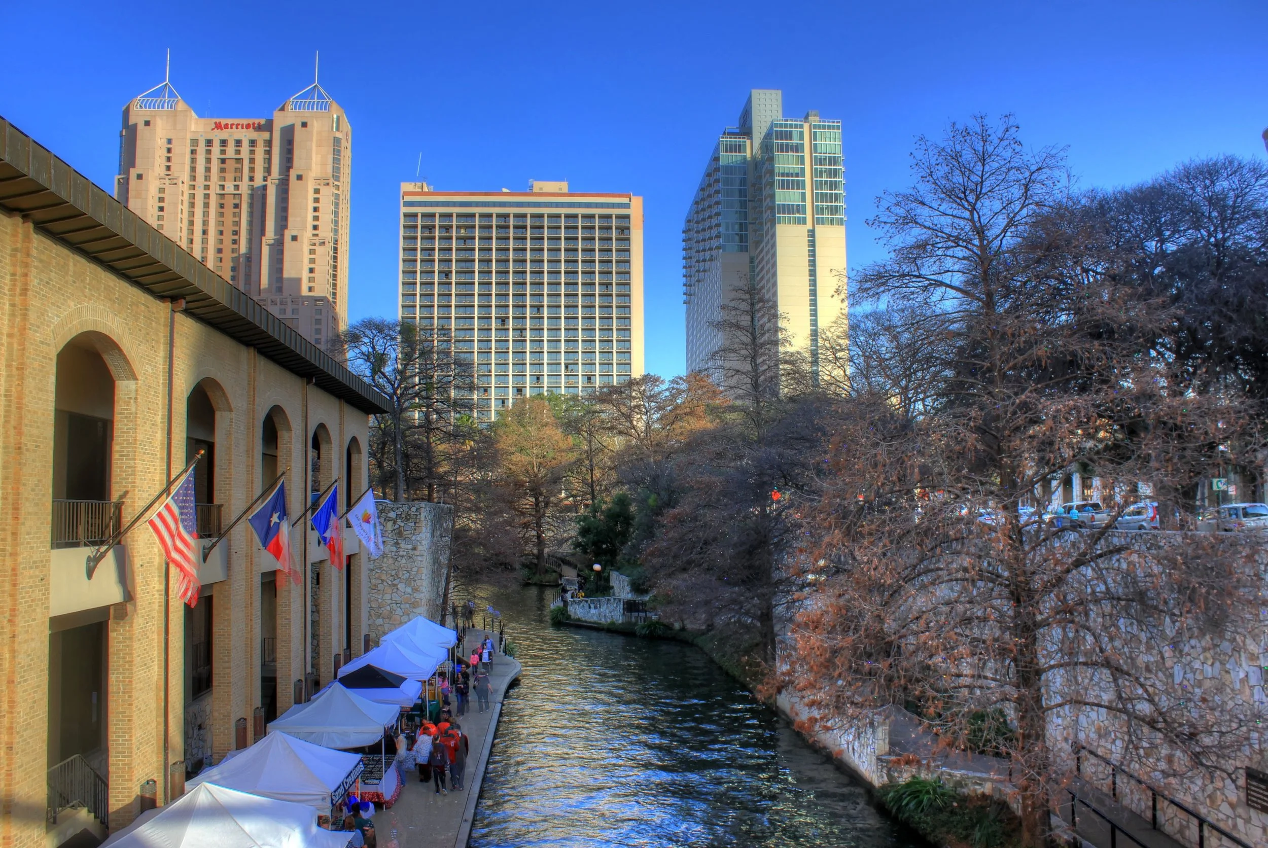 Walkway beside a canal lined with white tents, with hotel towers, flags and bare trees behind