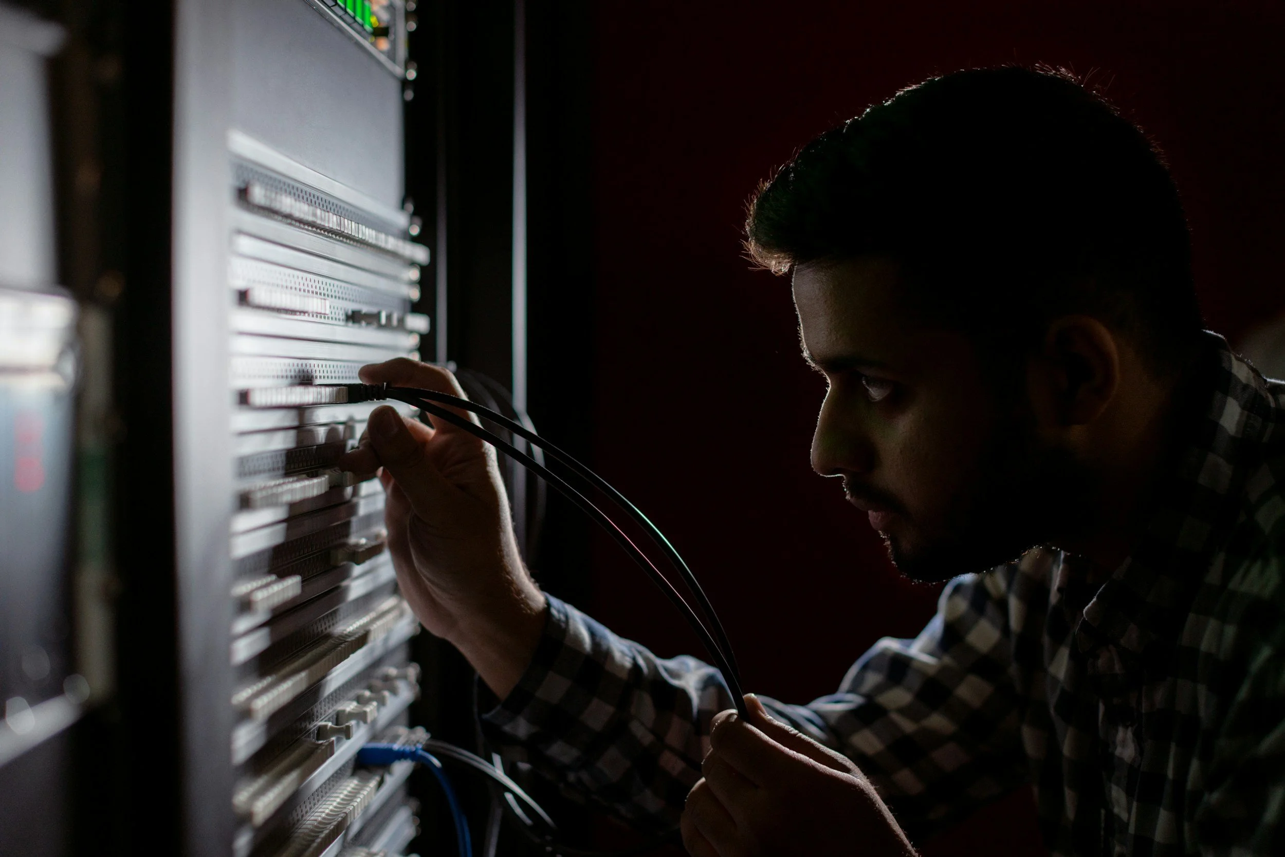 A man in a plaid shirt plugs network cables into a rack in a dim server room