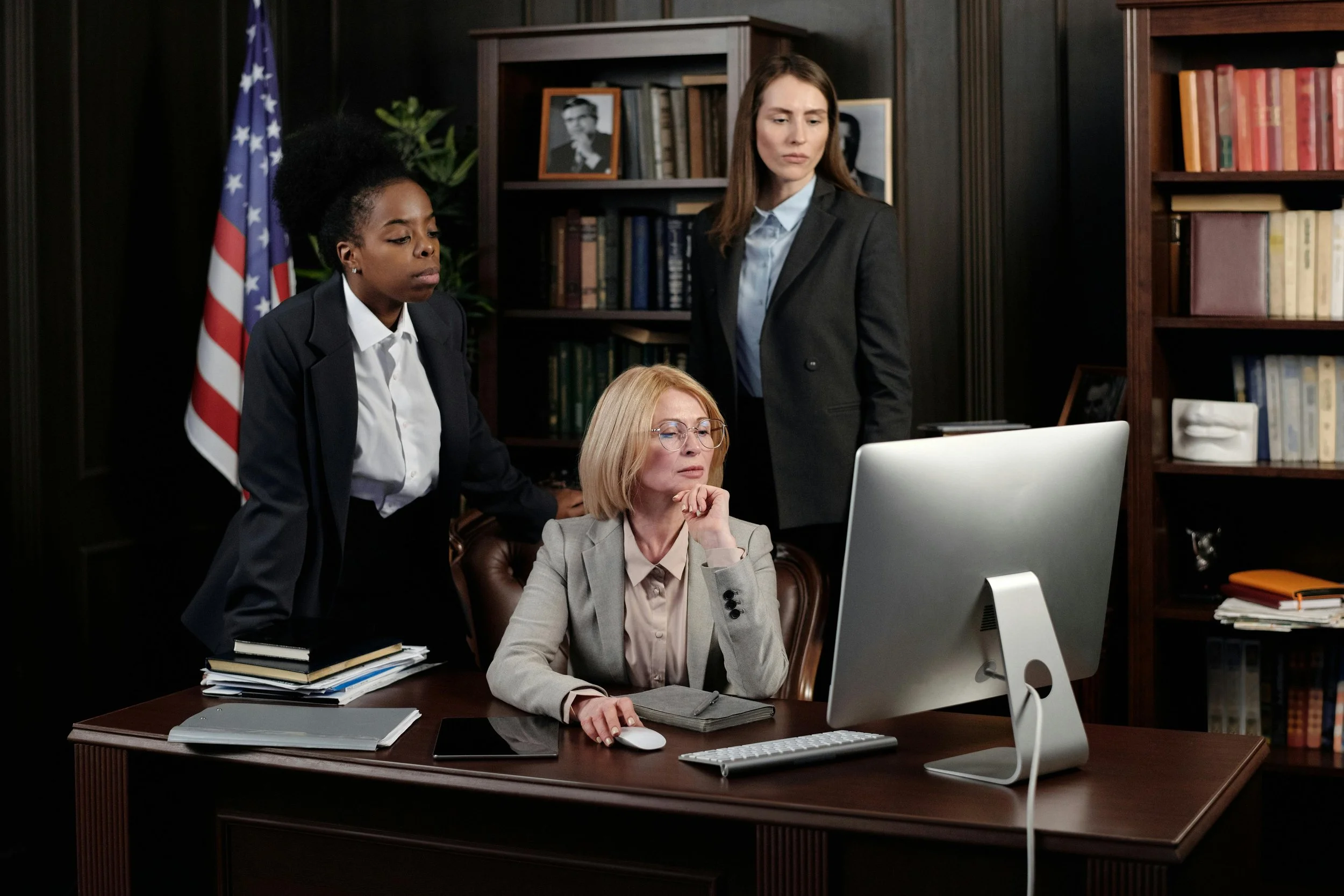 Three women in dark business attire gather around a desktop computer in a wood-paneled office with an American flag