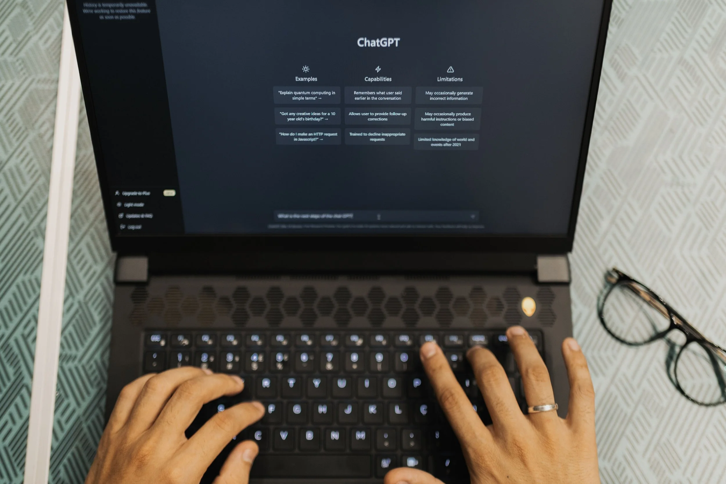 Hands typing on a laptop that shows the ChatGPT start screen, with eyeglasses resting on the desk beside it