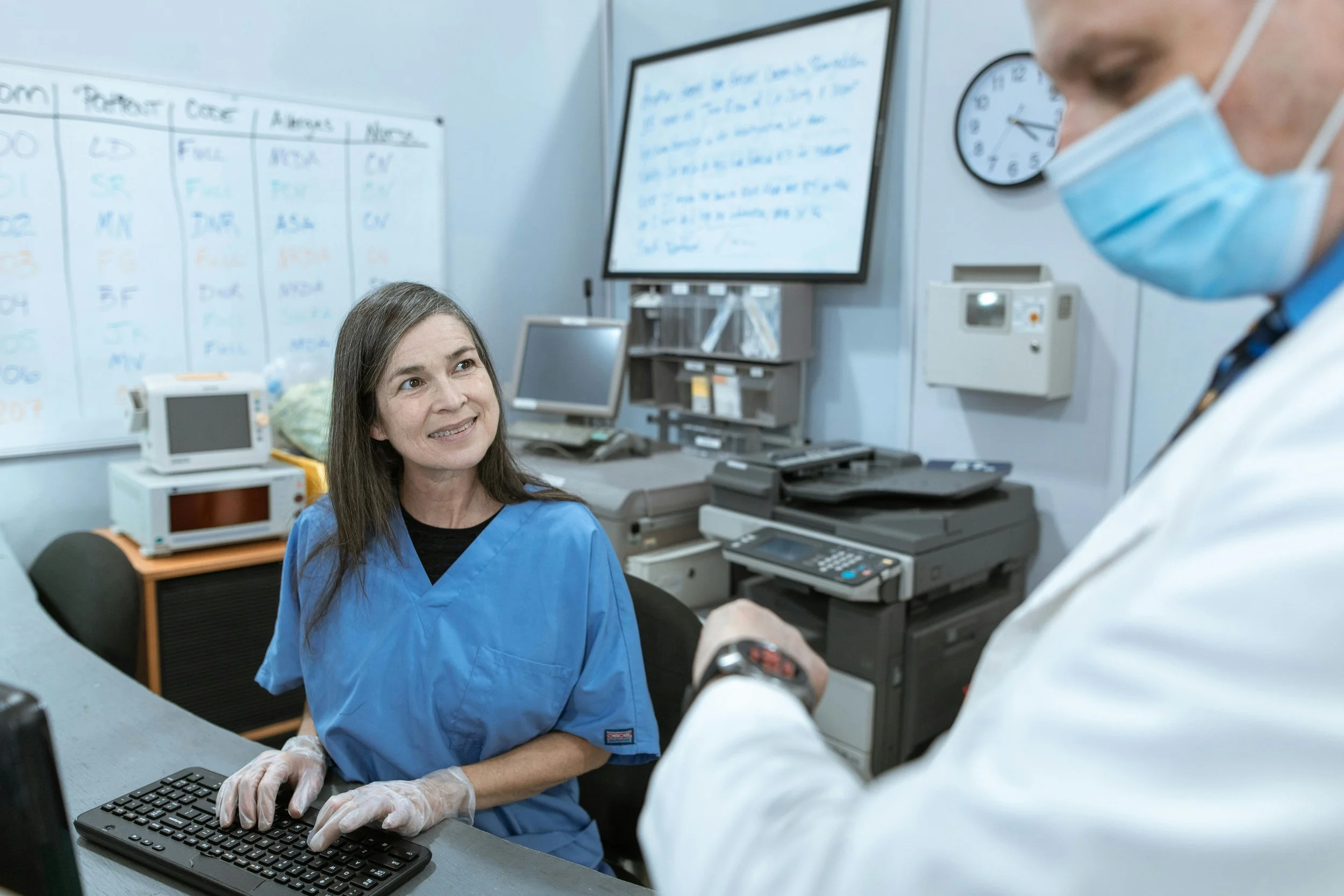 A nurse at a clinic workstation looks up at a doctor in a mask standing beside her